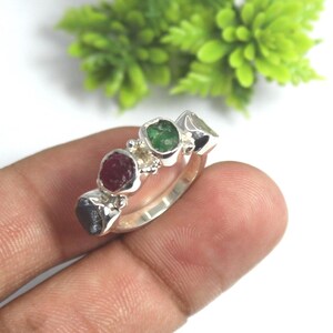 Peut inclure: Une bague en argent ornée de quatre pierres précieuses colorées : un rouge profond, un vert vif et deux pierres gris foncé. Le corps de la bague est lisse et les pierres sont serties dans un design en grappe. Le fond est flou avec un feuillage vert.