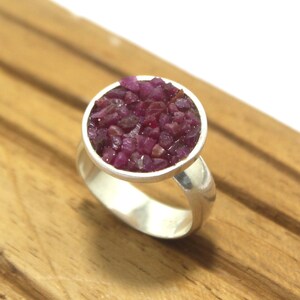 May include: A silver ring featuring a circular setting filled with small, deep red gemstones. The ring's band is smooth and polished, and the gemstones are clustered together, creating a textured surface. The ring is displayed on a wooden surface.
