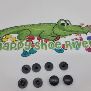 May include: Six black plastic shoe rivets. The rivets have a flat top and a small post on the bottom. The rivets are arranged in a semi-circle on a white surface. The image also includes a cartoon alligator with the text "snappy shoe rivets".