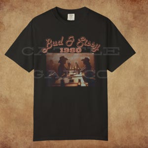 May include: Black t-shirt featuring the text "Bud & Sissy 1980" in a retro font. The shirt has a graphic of two people in cowboy hats. The shirt is made of a soft material and is perfect for casual wear.