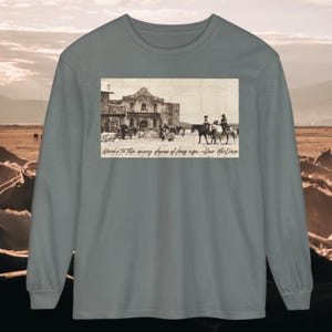Op de afbeelding: Een lange mouwen shirt in een gedempte teal kleur. Het shirt heeft een sepia-getinte afbeelding van een historisch gebouw met mensen en paarden, en de tekst "Here's to the many dupes of long ago. - Gus McCrae."