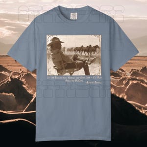 May include: Dusty blue t-shirt featuring a sepia-toned image of a man in a cowboy hat, seated in a chair, with horses in the background. The text reads "Let the English play Hamlet and King Lear - I'll play Augustus McCrae - Robert Duvall."