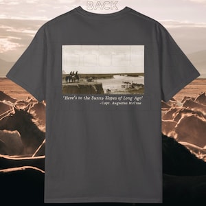 May include: Dark gray t-shirt featuring a sepia-toned image of people on horseback near a river. The text "Here's to the Sunny Slopes of Long Ago" and "-Capt. Augustus McCrae" are printed below the image. The word "BACK" is printed at the top.