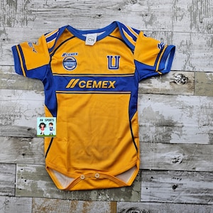 May include: A yellow and blue baby bodysuit with the text "CEMEX" and the letter "U". The bodysuit has short sleeves and a round neckline. The design includes the team logo and the word "Monterrey".