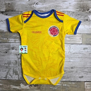 May include: Yellow baby bodysuit with blue and red accents, featuring the word "COLOMBIA" and a soccer ball emblem. The bodysuit has short sleeves and a snap closure. The design is inspired by the Colombian national soccer team.