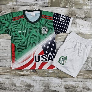May include: A green and white soccer jersey with the words "Mexico" and "USA" printed on it. The jersey has a red, white, and blue American flag design on one side and a green and white geometric pattern on the other side. The jersey is paired with white soccer shorts with a green logo.