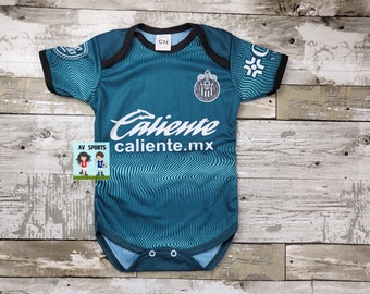 May include: A teal blue baby onesie with a white "Caliente" logo and a team logo on the front. The onesie has a black trim and a white wave pattern.