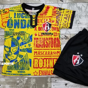 May include: A yellow and black soccer jersey with a wrestling mask design and the text "LUCHA" and "URREA". The jersey is paired with black soccer shorts with a red and white logo.