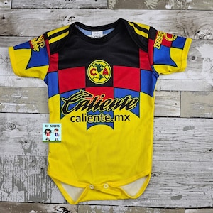 May include: Yellow, black, red, and blue baby bodysuit with the text "Caliente" and "caliente.mx". The bodysuit has a soccer team logo on the chest and a checkered pattern. The sleeves have yellow and blue accents.