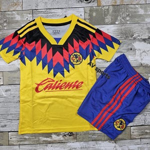 May include: A yellow soccer jersey and blue shorts set. The jersey has a V-neck and a geometric design in red, blue, and black. The word "Caliente" is printed in red. The shorts are blue with red stripes.