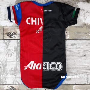 May include: Red and black baby bodysuit with the text "CHIVAS" and "Akiko" in white. The bodysuit has blue trim and the text "Coca Cola" and "&eacute;xico" on the sleeves.