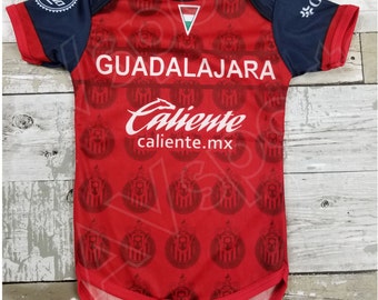 May include: Red and navy blue baby onesie with a white logo that says "Guadalajara Caliente caliente.mx". The onesie has a repeating pattern of a soccer team logo.
