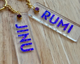 May include: Two clear acrylic keychains with gold-colored hardware. One keychain reads "JINU" in blue and pink glitter lettering, and the other reads "RUMI" in the same style. Each keychain has a small purple gem attached.