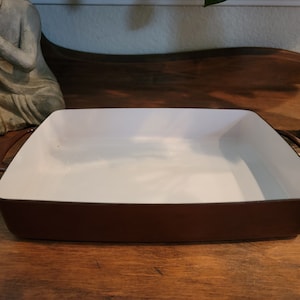 Puede incluir: Una fuente de horno cuadrada con interior blanco y exterior marrón. La fuente tiene dos asas marrones y está sobre una superficie de madera. La fuente está vacía.