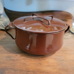 Puede incluir: Una olla de hierro fundido esmaltada marrón con tapa y asa de metal. La olla está sobre una superficie de madera.