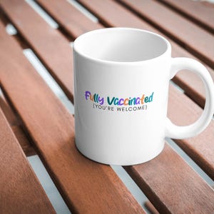 May include: White ceramic mug with the text "Fully Vaccinated (you're welcome)" in colorful lettering. The mug has a curved handle and sits on a wooden surface.