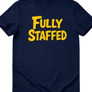May include: Navy blue t-shirt with the text "Fully Staffed" in yellow, bold, block letters. The shirt is a crew neck style and appears to be made of a soft material. The text is centered on the front of the shirt.