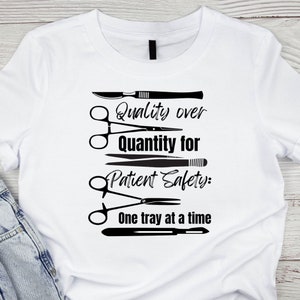 May include: White t-shirt with a black graphic design of surgical instruments and the text "Quality over quantity for Patient Safety: One tray at a time."