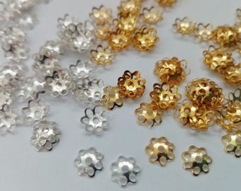 以下が含まれることがあります： 金と銀の金属製のビーズキャップのコレクション。キャップは花の形をしていて、中央に丸い穴があります。キャップは白い表面に散らばっています。
