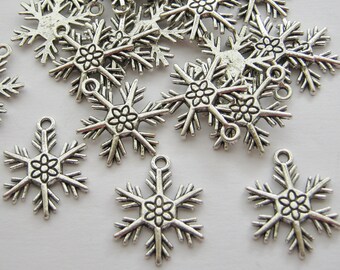 May include: A collection of silver-colored snowflake charms. Each charm features a six-pointed design with a flower in the center and a small loop at the top for stringing. The charms have a vintage, slightly tarnished appearance.