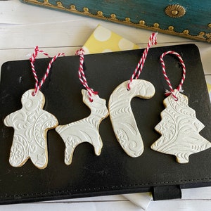Puede incluir: Cuatro adornos navideños blancos con diseños en relieve y bordes dorados. Los adornos incluyen un hombre de jengibre, un reno, un bastón de caramelo y un árbol de Navidad, cada uno con una cuerda roja y blanca. Sobre una superficie negra.