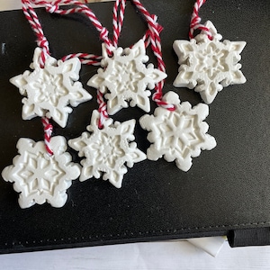 Puede incluir: Seis adornos blancos con forma de copo de nieve con diseños intrincados. Cada adorno está suspendido por una cuerda de hilo rojo y blanco. Los adornos están dispuestos sobre una superficie negra, lo que sugiere un tema navideño o invernal.