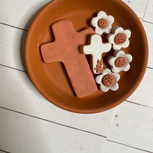 Op de afbeelding: Een terracotta schaal met een wit kruis en vijf witte bloemen met bruine centra. Het kruis en de bloemen zijn van keramiek.