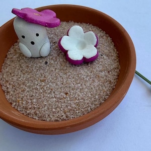 Puede incluir: Un cuenco de color terracota lleno de arena, con una pequeña figura en forma de hongo blanco con una gorra de flor rosa y una flor blanca y rosa. Una ramita de lavanda es visible a la derecha.