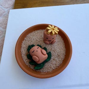 May include: A small terracotta pot filled with sand, featuring two miniature clay figures. One figure is upright with a flower on its head, and the other is lying down on green leaves. The figures are peach-colored with simple facial features.
