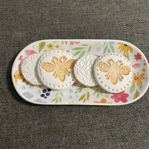 May include: An oval ceramic serving tray with a floral design, holding four white coasters. Two coasters feature a gold bee design, and two have a floral relief pattern. The tray is oval-shaped.