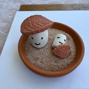 May include: A terracotta-colored ceramic bowl filled with sand, featuring a whimsical mushroom and leaf design. The mushroom has a white stem and a brown cap with leaf detailing. A smaller leaf and mushroom are also present.