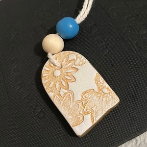 May include: A cream-colored, floral-patterned pendant with a beige border, strung on a white cord with a light blue and beige wooden bead. The pendant has a rectangular shape with rounded edges.