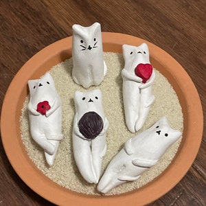 Könnte beinhalten: Fünf weiße Keramik-Katzenfiguren mit bemalten Details, die in einer Terrakotta-Schale mit Sand sitzen. Einige Katzen halten kleine rote und lila Herzen.