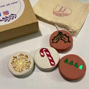 May include: A set of four round, decorative Christmas ornaments. One ornament features a gold snowflake, another a red and white candy cane, a third a green holly design, and the last has three green Christmas trees. The ornaments are in a gift box and a small bag.
