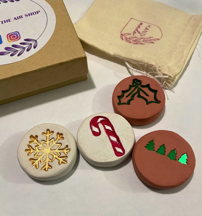 May include: A set of four round, decorative Christmas ornaments. One ornament features a gold snowflake, another a red and white candy cane, a third a green holly design, and the last has three green Christmas trees. The ornaments are in a gift box and a small bag.