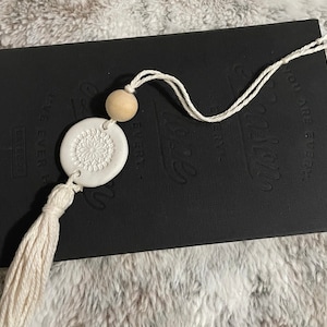 Puede incluir: Marcapáginas de cerámica blanca con un diseño de mandala detallado, un borla blanca y una cuenta de madera. El marcapáginas está unido a un diario negro con el texto "Every Story Matters" en relieve en la portada.