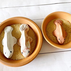 May include: Two white ceramic fish and one brown ceramic fish are displayed in terracotta bowls filled with sand. The fish are all facing the same direction.