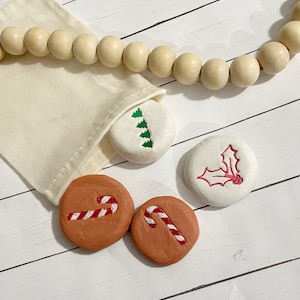 May include: A set of painted Christmas-themed stones. The stones feature designs of candy canes, a Christmas tree, and holly. They are in a small beige bag, with a wooden bead garland in the background. The stones are on a white wooden surface.