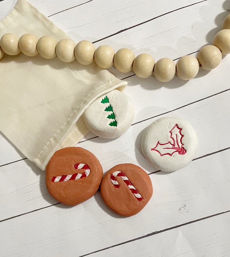 May include: A set of painted Christmas-themed stones. The stones feature designs of candy canes, a Christmas tree, and holly. They are in a small beige bag, with a wooden bead garland in the background. The stones are on a white wooden surface.