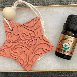 May include: A terracotta star-shaped ornament with a floral design, a wooden bead, and a string for hanging. A small bottle of USDA Organic Lavender essential oil is included in the image.