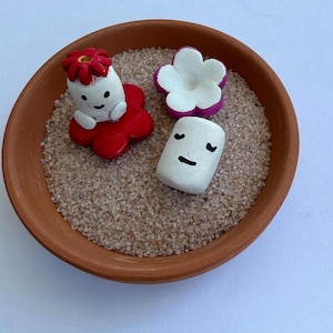 May include: A terracotta bowl filled with sand, containing three small, hand-painted ceramic figures. One figure is white with a red flower, another is a white flower with purple edges, and the third is a white cylinder with a simple face.