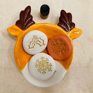 May include: A ceramic reindeer-shaped dish in yellow, brown, and white, holding three decorative stones. Two white stones feature gold holly and snowflake designs, and one orange stone has a gold snowflake. A small brown bottle sits above the dish.