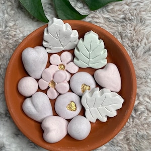 May include: A collection of small, decorative, pastel-colored clay ornaments in the shape of hearts, leaves, and flowers. The ornaments are arranged in a terracotta bowl.