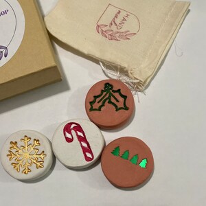 May include: A collection of four round, decorative Christmas ornaments. One ornament features a gold snowflake, another a red and white candy cane, and two others have green holly and Christmas tree designs. The ornaments are displayed with a small gift box and a fabric bag.