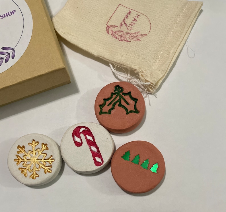 May include: A collection of four round, decorative Christmas ornaments. One ornament features a gold snowflake, another a red and white candy cane, and two others have green holly and Christmas tree designs. The ornaments are displayed with a small gift box and a fabric bag.
