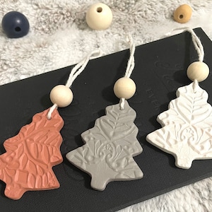 Puede incluir: Tres adornos navideños con forma de árbol en tonos terracota, gris y blanco. Cada árbol tiene un diseño floral en relieve detallado y una cuenta de madera en la parte superior, sujeta a una cuerda blanca para colgar. Los adornos están sobre una superficie oscura.
