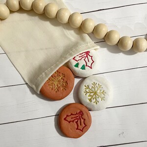 May include: A beige fabric bag with wooden beads and several round, terracotta-colored and white stones. The stones feature embossed holiday designs, including snowflakes and holly. The bag is on a white wooden surface.