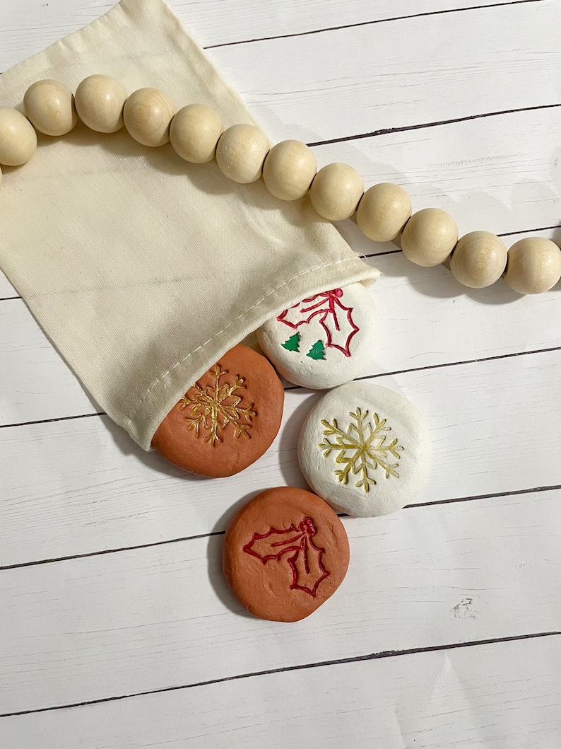 May include: A beige fabric bag with wooden beads and several round, terracotta-colored and white stones. The stones feature embossed holiday designs, including snowflakes and holly. The bag is on a white wooden surface.
