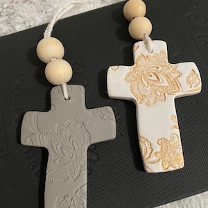 May include: Two ceramic cross ornaments with a floral design. The crosses are white and grey with gold accents. The crosses are hanging from a string with wooden beads.