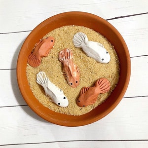 Op de afbeelding: Vijf keramische visfiguren in een terracotta kom gevuld met zand. De vissen zijn geschilderd in tinten oranje en wit.